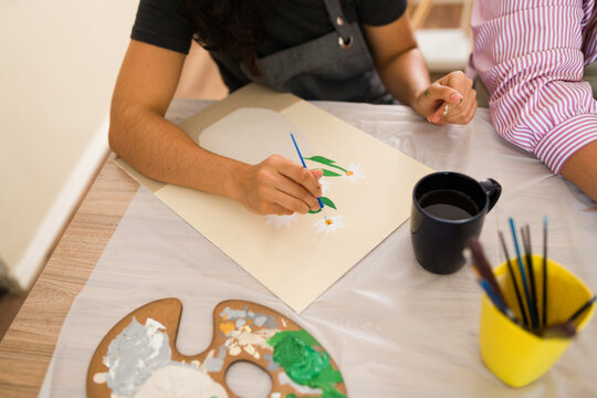 Woman's hand holding paintbrush creating flower artwork during art class, learning creative hobby with friends