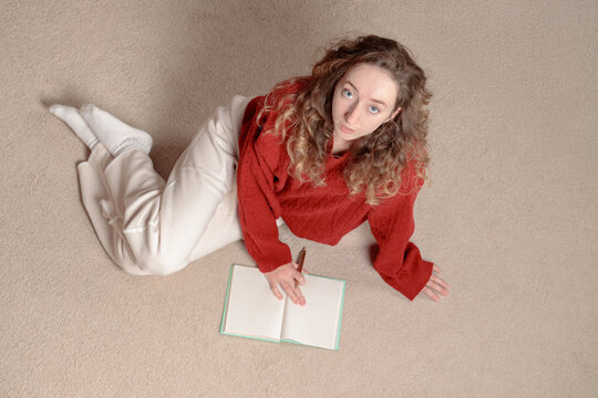 teenage girl with curly hair sitting on the floor and writing in her notebook at home