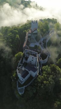 Static top-down aerial view of Celje Castle in Slovenia, showing medieval walls, courtyards and historic buildings surrounded by forest and drifting fog.
