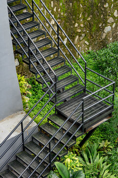 Black industrial metal staircase with grated steps and railings beside a moss-covered stone wall, surrounded by lush green tropical plants.