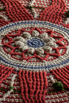 Close-up of colorful handmade crochet textile with intricate circular floral pattern, textured yarn, raised stitches, and red, beige, blue and green details.