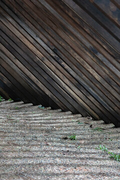 Abstract architectural detail of dark weathered wooden slats arranged diagonally above textured ground, creating strong geometric lines and repeating patterns.