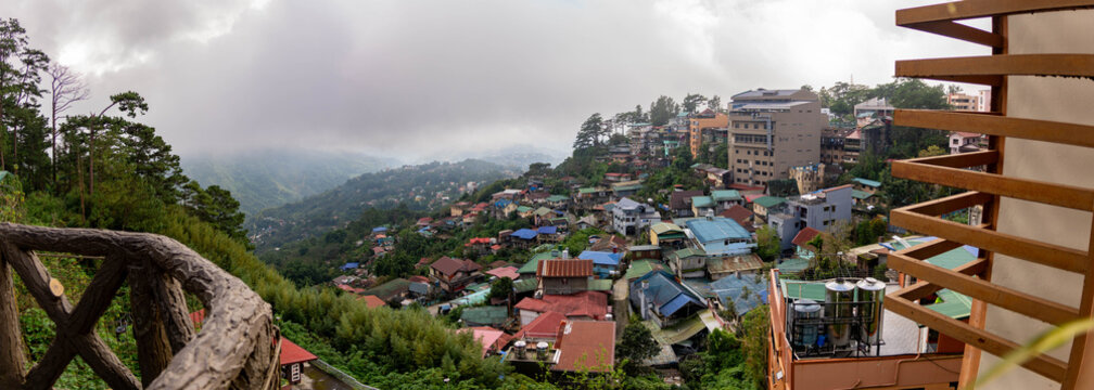 Panoramic view of a densely built mountain city surrounded by lush green hills under low clouds and mist, showing colorful hillside homes and urban development.