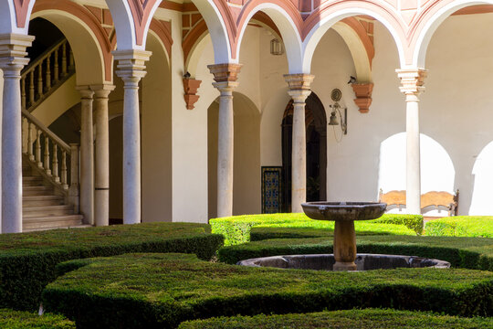 Mudejar style architecture creating a serene courtyard garden, featuring lush green hedges, ancient arches, elegant pillars, and a historical stone fountain under natural sunlight