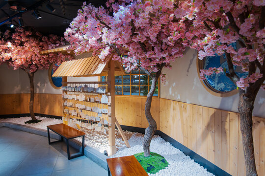 Japanese shrine interior decoration with blooming pink cherry blossom trees, wooden ema wishing plaque stand, and white gravel zen garden.