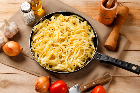 Cooked fettuccine served in a black skillet on a wooden table, accompanied by garlic, onion, tomato, olive oil, and salt.