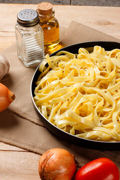 Cooked fettuccine pasta presented in a black pot on a wooden table, surrounded by garlic, onion, tomato, olive oil, and salt.