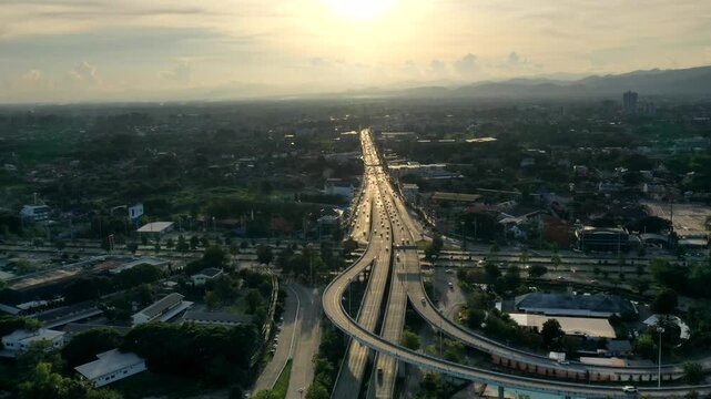 Aerial hyperlapse pan over Don Chan Intersection in Chiang Mai, Thailand, revealing the multilane highways, curved ramps and suburban neighborhoods stretching toward distant mountains at radiant dawn.