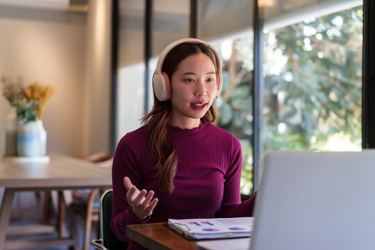 Young Asian businesswoman wearing headphones talks during video call with laptop in modern office