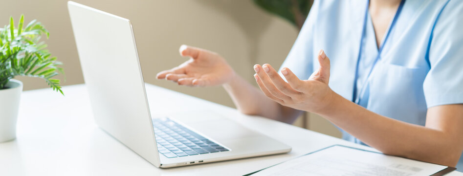 Professional Asian female doctor or nurse in blue uniform gesturing during online telehealth medical consultation with patient on laptop computer in office with medical chart on table.