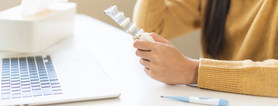 Asian young woman holding a blister pack of pills during an online medical consultation with laptop and thermometer on table, home healthcare and telemedicine concept.