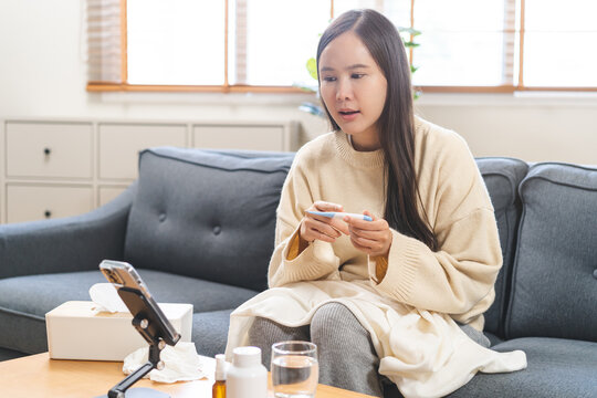 Sick young Asian woman holding digital thermometer showing temperature during video call online consultation with doctor via smartphone app on sofa at home