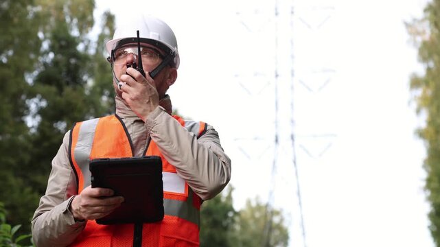 Energy engineer speaking on walkie talkie and holding tablet in protective case near power line. Using AI algorithms to optimise data, control smart grid work, and accelerate sustainable power.