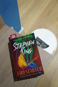 Stephen King Firestarter book cover resting on a wooden shelf near a white robotic vacuum cleaner