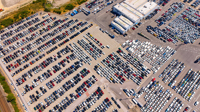 Vehicle storage blocks around warehouse roofs, aerial view. Cars occupy several paved sections divided by lanes, with industrial buildings near the upper edge.