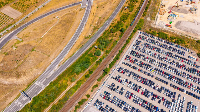 Vehicle storage beside road ramps and railway, aerial view. Parked cars fill the lower right corner, with tracks and asphalt lanes crossing dry grass.