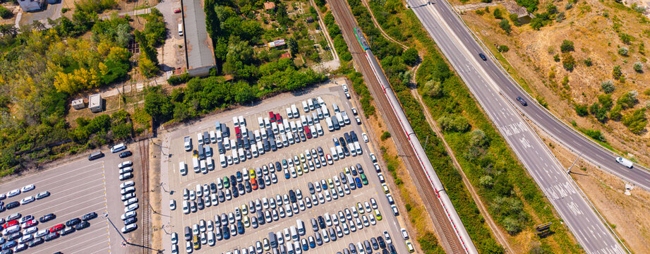 Aerial view of a passenger train moving past a car lot and highway. A red and white train travels along tracks between a parking lot filled with cars and a paved road.