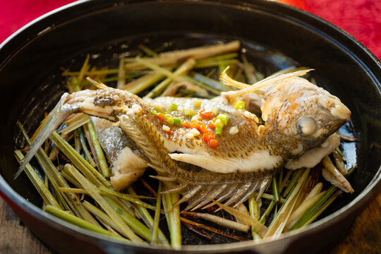 angle view of steamed yellowfin seabream on lemongrass in black plate garnished with chopped scallion and red chili