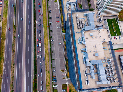 Highway traffic alongside office complex, aerial view. A queue of cars follows a service road beside rooftop machinery, parking spaces and planted terraces.