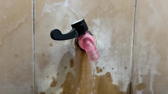 Old pink faucet handle dripping water intermittently on a stained tiled wall.