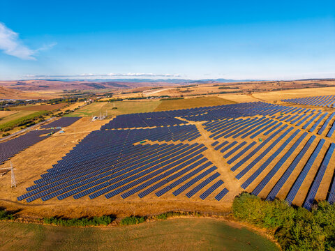 Photovoltaic field with diagonal panel rows, aerial view. Blue arrays occupy dry grass above a cultivated foreground, with hills along the horizon.