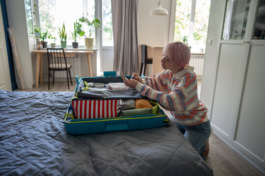 Thoughtful woman preparing for upcoming travel and writing checklist while sitting beside open case, relaxed female organizing personal luggage, packing suitcase and write list of items in notepad.