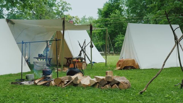 A serene historical reenactment camp with white tents, crackling campfire, and cooking pots in a lush green meadow, offering a glimpse into a bygone era.