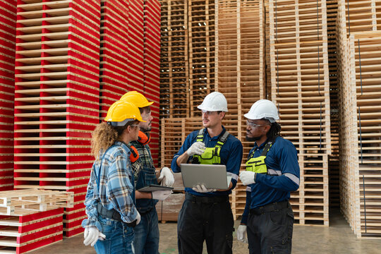 During factory teamwork, four workers hold a team meeting as a man holding a laptop addresses three colleagues beside wooden pallets.