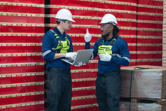 During lumber inventory checks, two warehouse colleagues discuss stock on a laptop beside tall stacks of sawn wood.