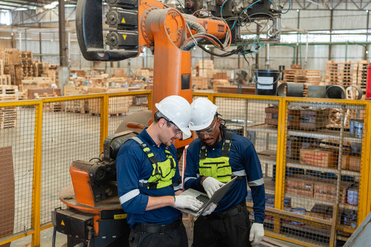 Reviewing robot automation during inspection, two men study a laptop beside the orange industrial robot arm inside a fenced cell.