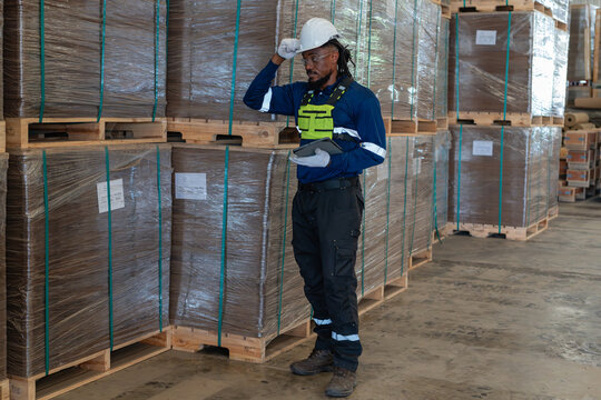 Among export pallets during packaging inspection, a man holds a tablet and touches his hard hat beside an export shipment.