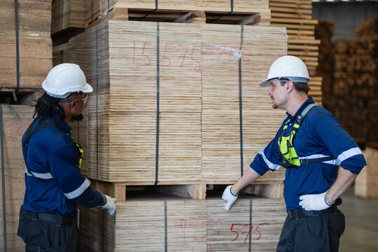Lumber inventory discussion: two warehouse colleagues look at each other beside stacked plywood resting on wooden pallets.