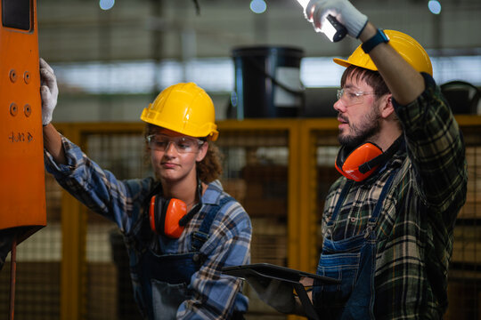 Industrial robot components undergo automation inspection as a man holds a tablet and flashlight beside a woman examining the orange arm.