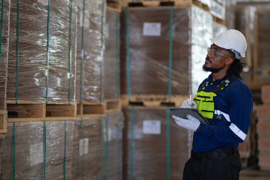 Export shipment packaging inspection: a warehouse worker holds a tablet while looking up at tall stacks of wrapped pallets.