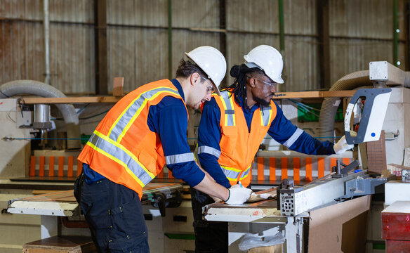 A carpenter documents plywood woodworking on a clipboard while his colleague operates the wood workshop panel saw control panel.