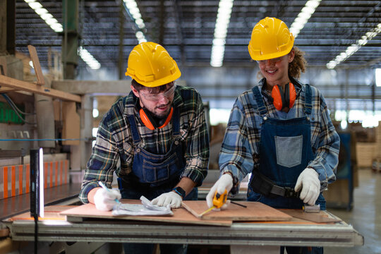A woman measures wood in the carpentry workshop; the female carpenter and her colleague take notes beside a panel saw.