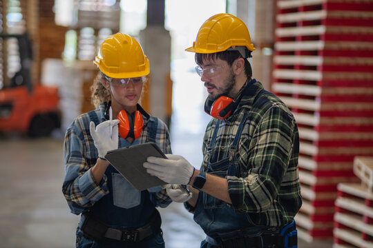 Inside the factory, two workers show teamwork as they review a tablet together beside tall stacks of wooden pallets.