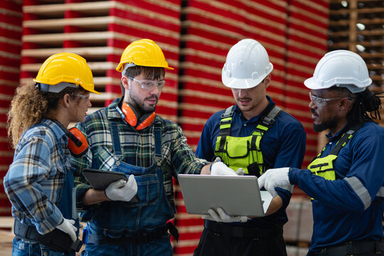 Discussing factory teamwork, four workers hold a team meeting while reviewing a laptop beside a tablet and stacked boards.