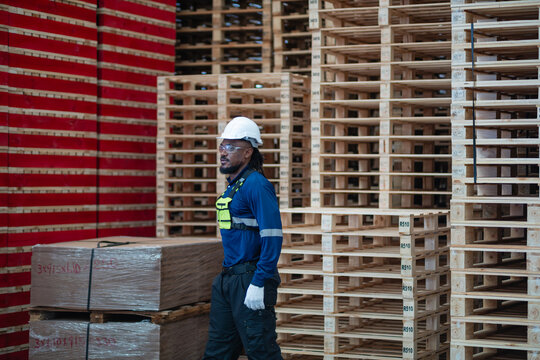 Wooden pallet stacks in a logistics warehouse surround a man walking past tall piles of pallets and wrapped lumber.