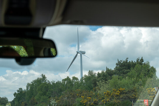A wind turbine rises above trees and yellow-flowering gorse, framed through a car's rear windscreen and mirror.
