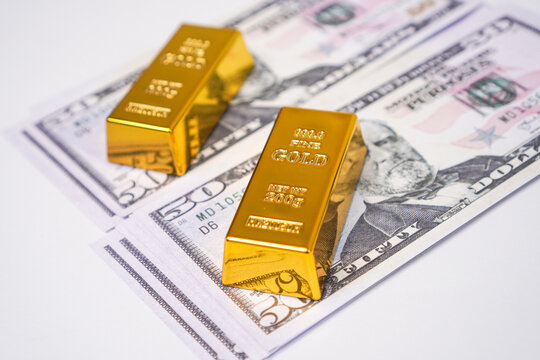 A high angle diagonal view of two shiny fine gold bullion bars placed over a scattered background of fifty dollar bills.