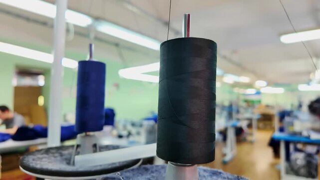 Black thread spool in sharp foreground of textile workshop, blue spool and fabric in background, blurred sewing tables, workers at work, and bright ceiling lighting with soft depth.