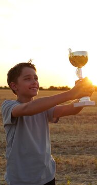 Happy boy holds trophy at sunset in rural field. Young winner celebrates victory and achievement outdoors. Golden hour sports success inspires pride and motivation for youth competition dreams.