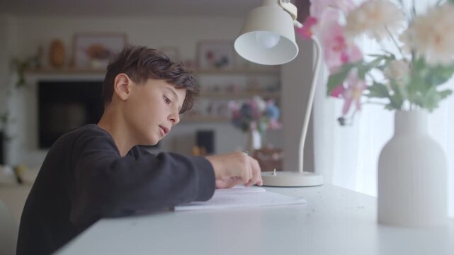 Focused young student writing with a pencil and using an eraser while sitting at a white desk in his room, concentrating on his studies and completing assignments for school at home