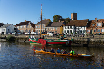 A team of rowers in a long wooden rowing boat practicing on the calm water of Ribe harbour basin in front of historic colourful townhouses. © Richard
