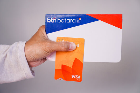 Makassar, Indonesia, September 20, A person's hand holding a BTN Batara iB sharia savings book along with an orange BTN Visa Gold debit card against a plain light gray background.
