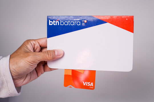 Makassar, Indonesia, September 20, A person's hand holding a BTN Batara iB sharia savings book along with an orange BTN Visa Gold debit card against a plain light gray background.