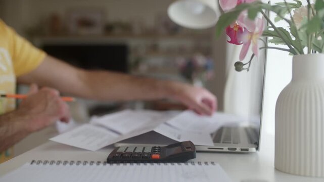 Man managing personal finances by calculating expenses and monthly bills with a calculator, writing notes in a notebook at his home office desk while organizing financial documents