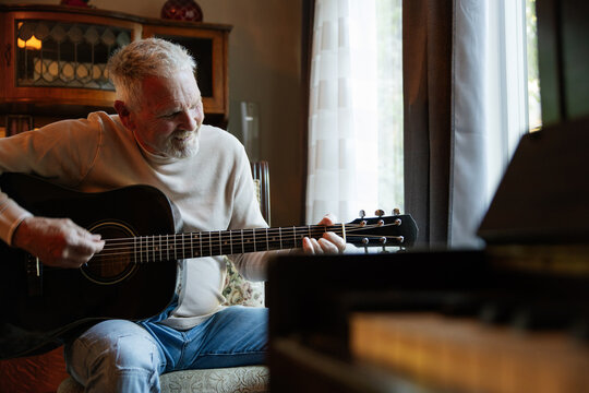 Older man playing acoustic guitar at home by a window