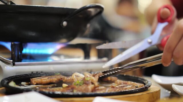 A close up cinematic shot of hands using tongs and scissors to cut and flip korean barbecue meat on a sizzling hot plate in a restaurant with a stove and boiling pot in the background.